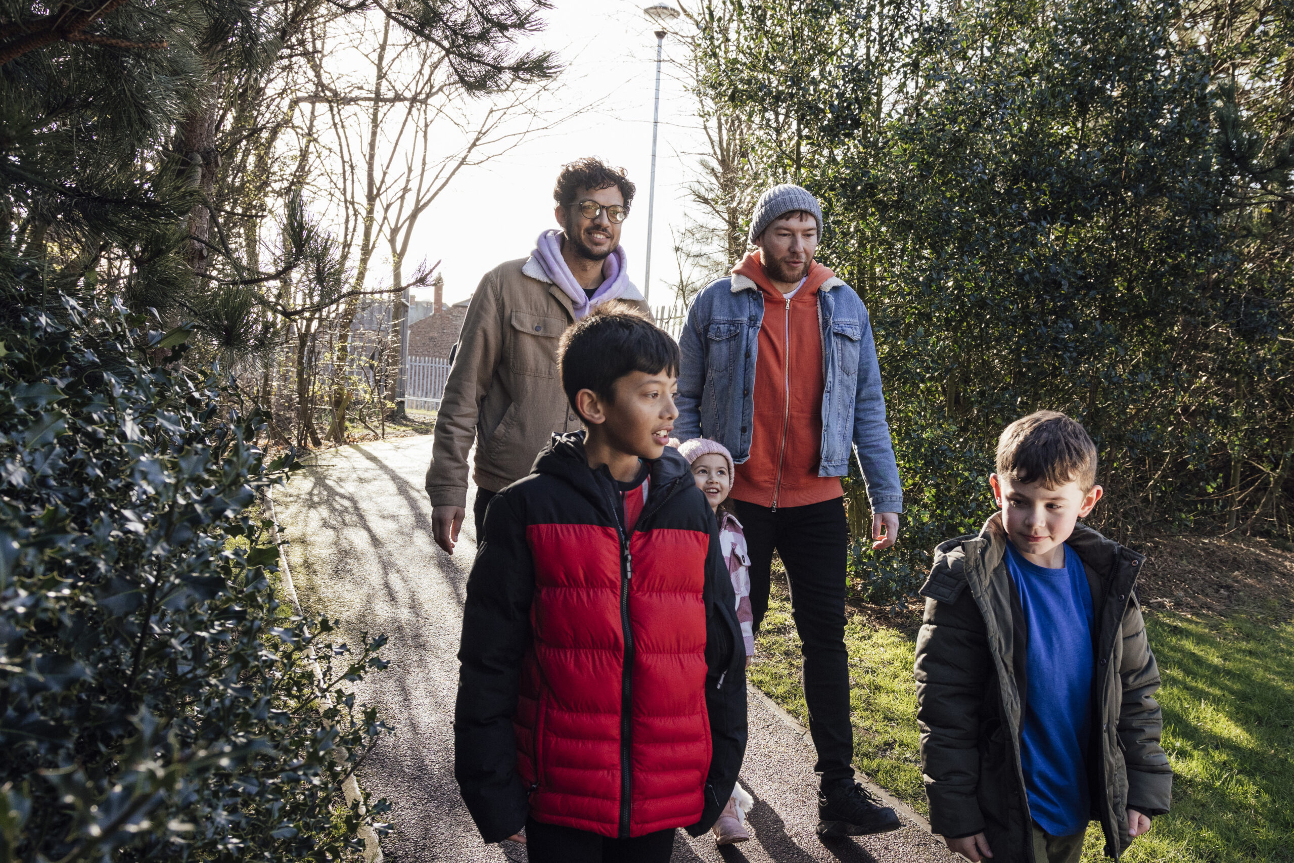 A wide-angle front view of a family of four a same-sex couple and their three foster children enjoying a day out in a park in the North East of England. They are all wrapped up warm for the cold weather. The two brothers are walking ahead while their sister hangs back with their foster carer