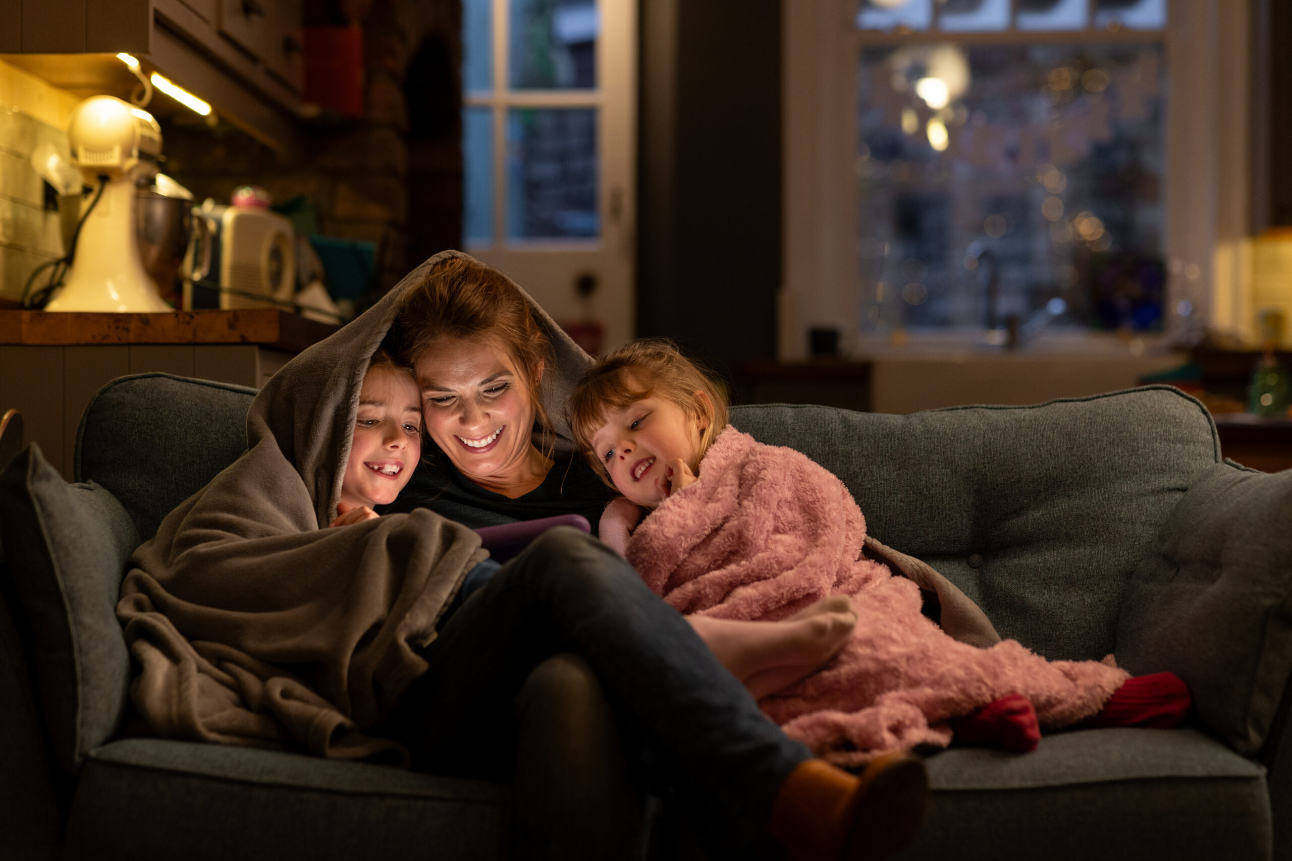 A shot of two young girls with their foster carer, sitting comfortably with a blanket over their heads on a sofa in the living room of their home in South Shields, North East England. They are all smiling, looking at a digital tablet which the foster parent is holding. In the background the kitchen is in partial view, and it is dusk outside.