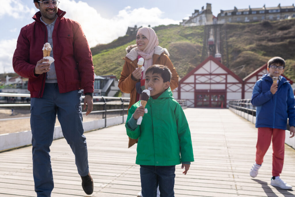 foster family visiting at beach on a staycation at Salburn by the Sea, North East of England. They are enjoying ice creams, wrapped up warm in the Great British weather while walking along a boardwalk.