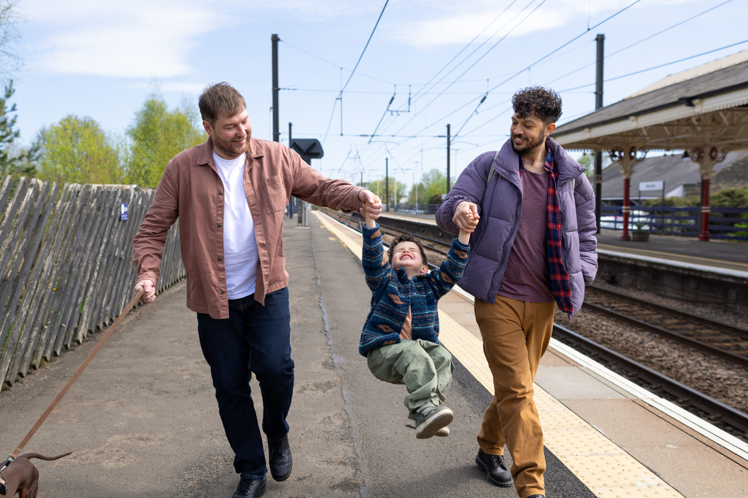 A full shot of a same-sex couple with their young son at Morpeth train station. Both parents lift the boy playfully, while their dachshund walks nearby. It's a sunny, calm day with train tracks in the background, capturing a candid family scene full of joy.
