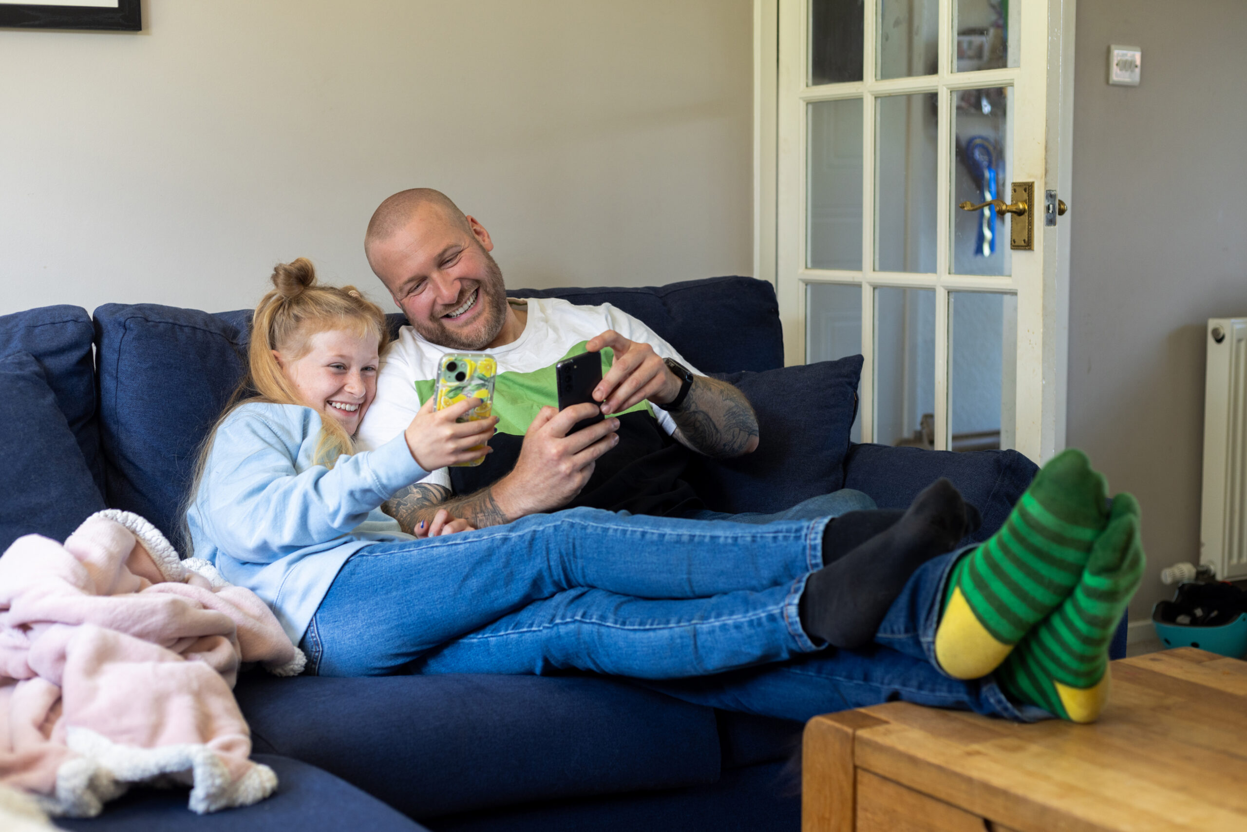 Medium shot of a girl and her male foster carer sitting together on a sofa in their family home in the North East of England. They are both holding mobile phones while smiling and looking at them. The male foster carer has his feet up on a table while the child has hers on the foster carer's legs.