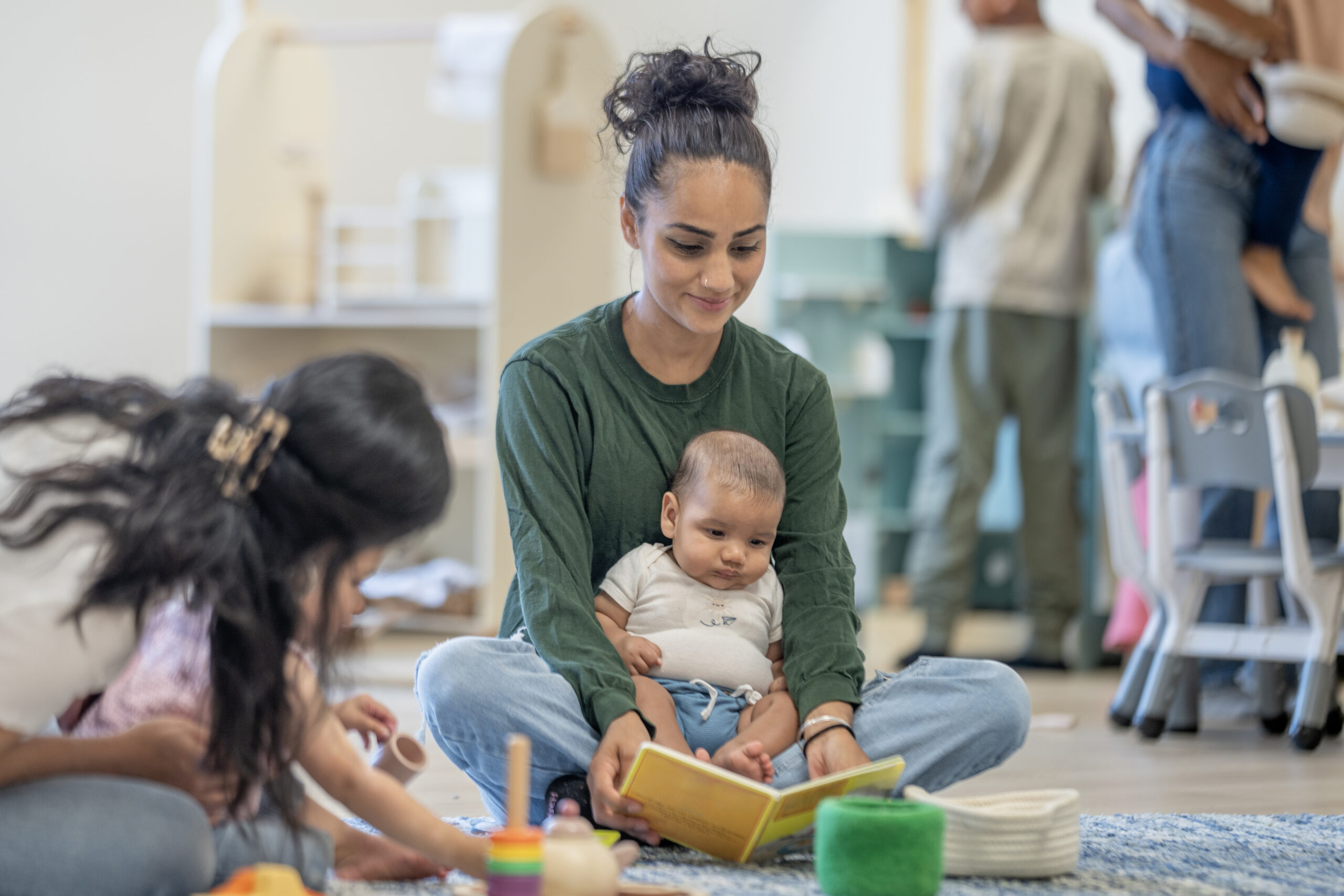A cheerful setting featuring a woman reading to a baby indoors, surrounded by children and parents engaging in various activities. The classroom promotes learning, bonding, and community.